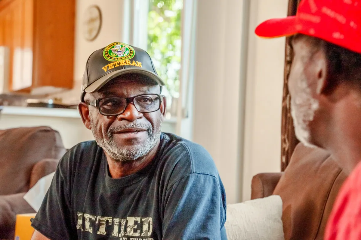 A smiling veteran in a veteran cap talking with a friend at home